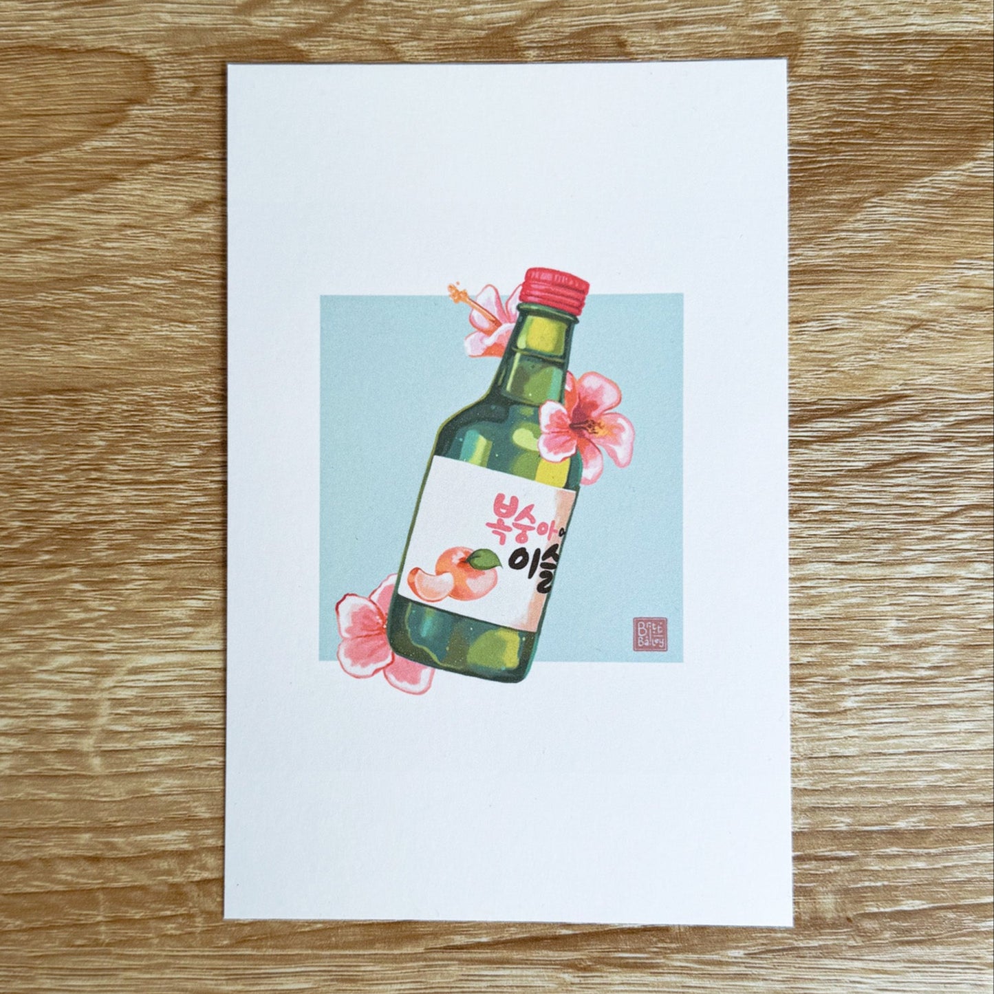 Hand holding a postcard featuring a painterly digital illustration of a bottle of peach flavoured korean soju surrounded by pink hibiscus flowers on a soft teal background.