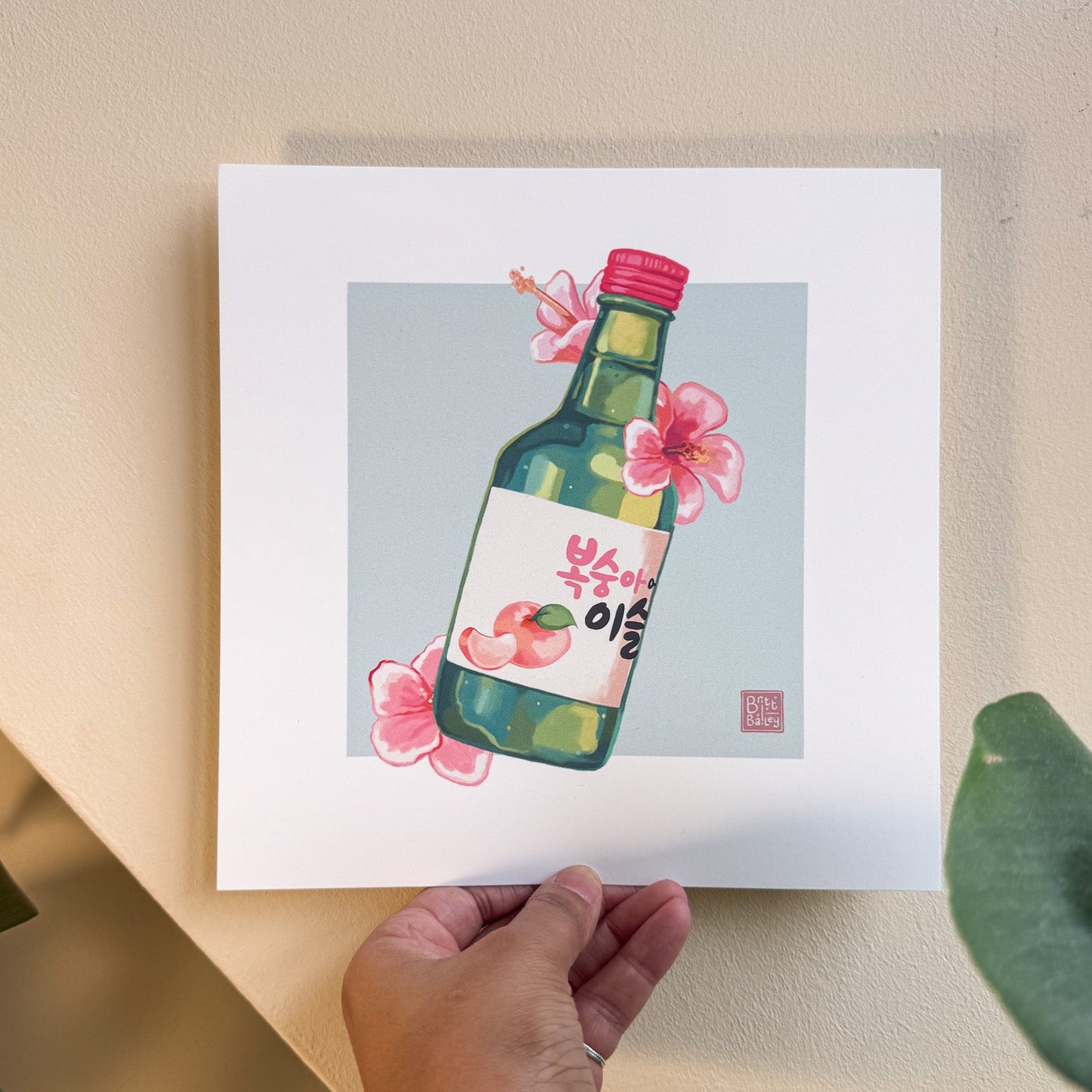 Hand holding an art print featuring a painterly digital illustration of a bottle of peach flavoured korean soju surrounded by pink hibiscus flowers on a soft teal background.