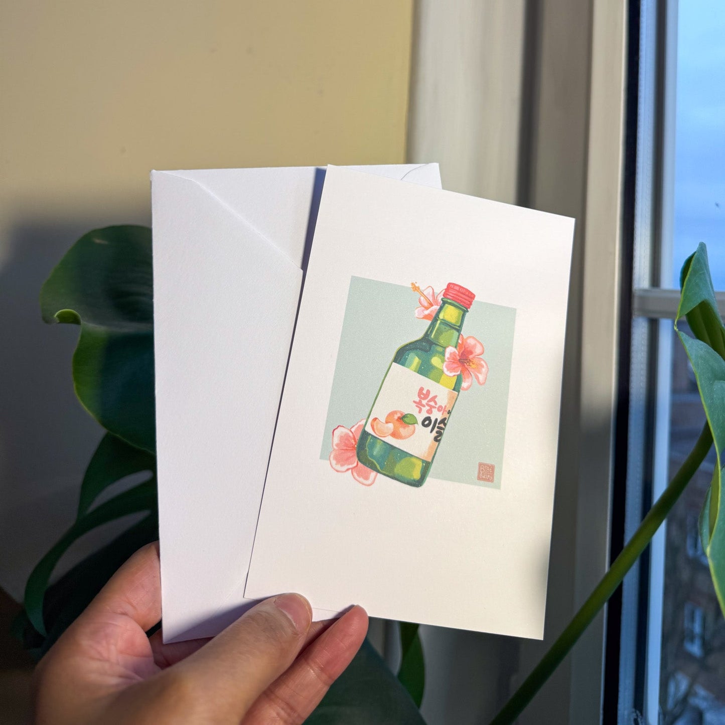 Hand holding a postcard featuring a painterly digital illustration of a bottle of peach flavoured korean soju surrounded by pink hibiscus flowers on a soft teal background.
