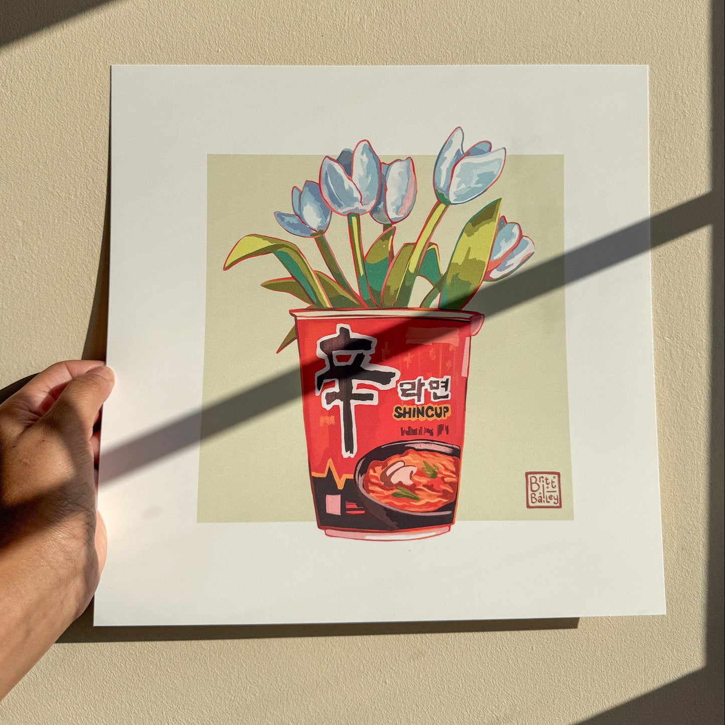 Hand holding an art print featuring a painterly digital illustration of cup of shin ramen noodles with a bouquet of white tulips inside on a soft green background.