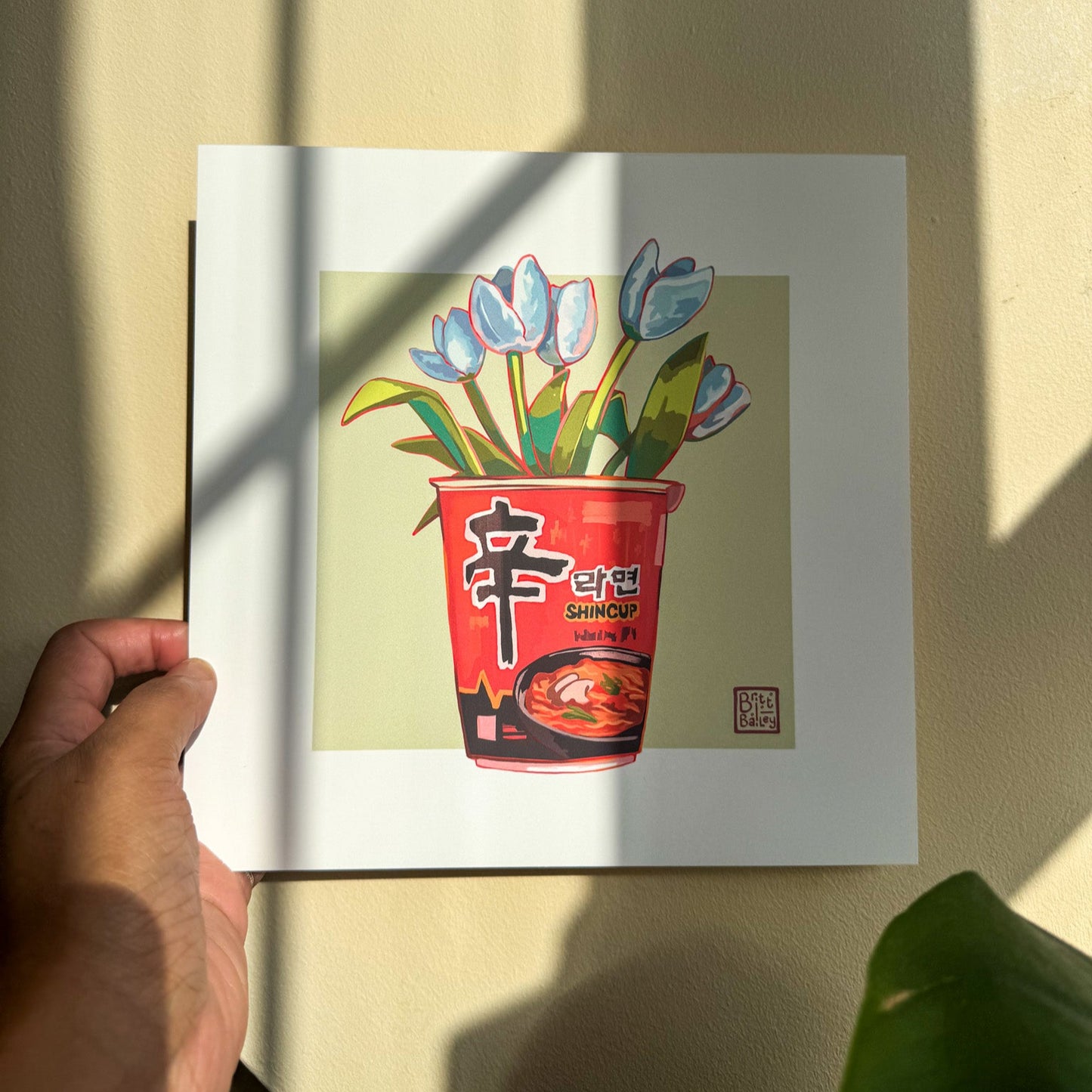 Hand holding an art print featuring a painterly digital illustration of cup of shin ramen noodles with a bouquet of white tulips inside on a soft green background.
