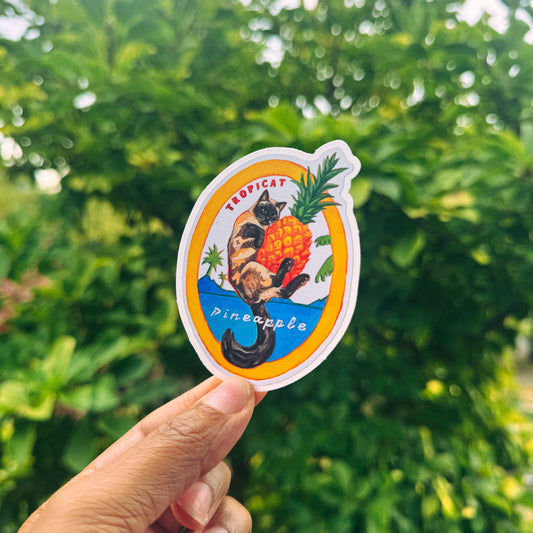 A hand holds a die-cut sticker featuring a tropical cat and pineapple design against a blurred green outdoor background.