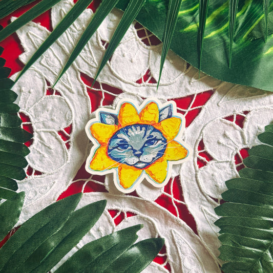 A die-cut sticker featuring a stylized cat face inside a yellow sunflower, displayed on a white lace textile with red background and green palm leaves.
