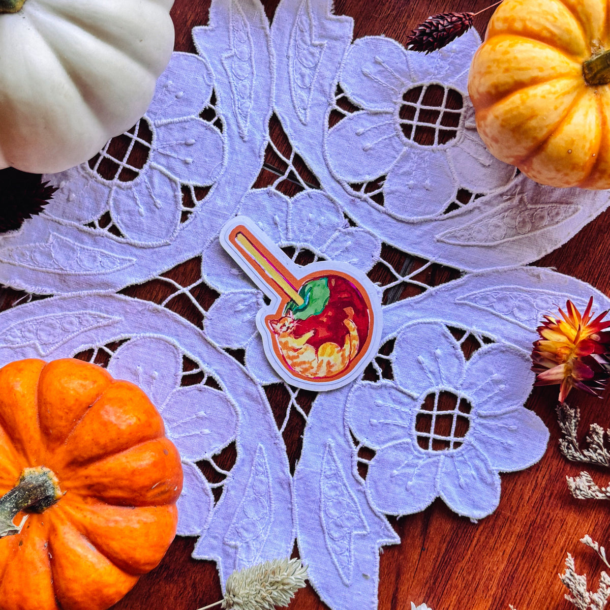 A decorative autumn flat-lay featuring a light purple lace doily with a small colorful sticker of a tan tabby cat in the center, surrounded by white and orange pumpkins, dried flowers, and a warm brown wooden surface.
