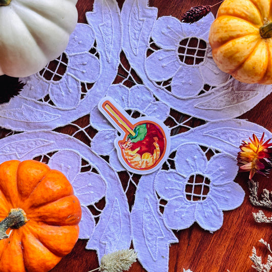A decorative autumn flat-lay featuring a light purple lace doily with a small colorful sticker of a tan tabby cat in the center, surrounded by white and orange pumpkins, dried flowers, and a warm brown wooden surface.