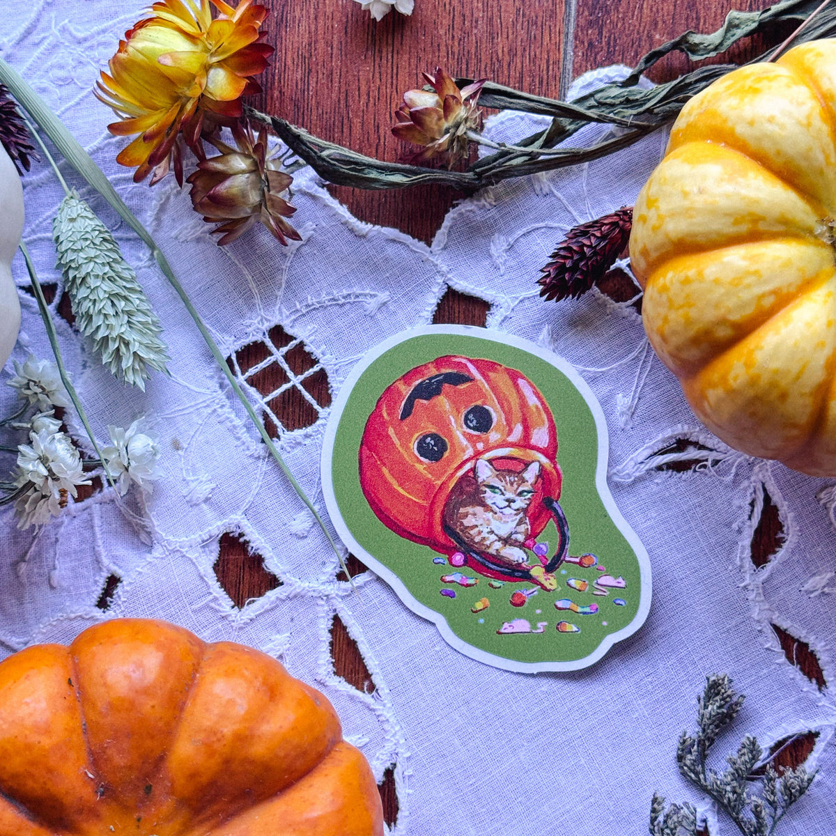 A die-cut sticker featuring a carved orange pumpkin with a cat inside, placed on a white lace cloth with autumn props including pumpkins, dried flowers, and pinecones.