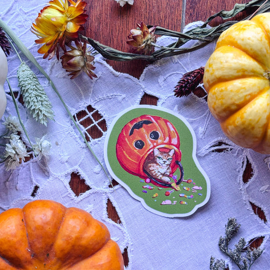 A die-cut sticker featuring a carved orange pumpkin with a cat inside, placed on a white lace cloth with autumn props including pumpkins, dried flowers, and pinecones.