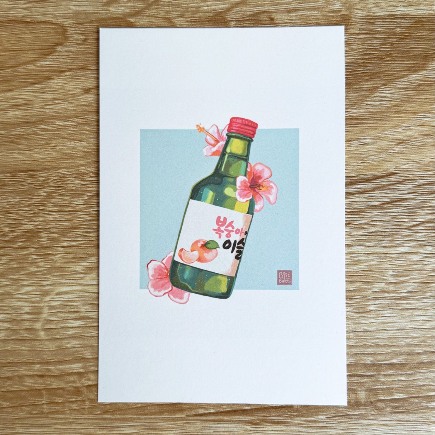 Hand holding a postcard featuring a painterly digital illustration of a bottle of peach flavoured korean soju surrounded by pink hibiscus flowers on a soft teal background. 