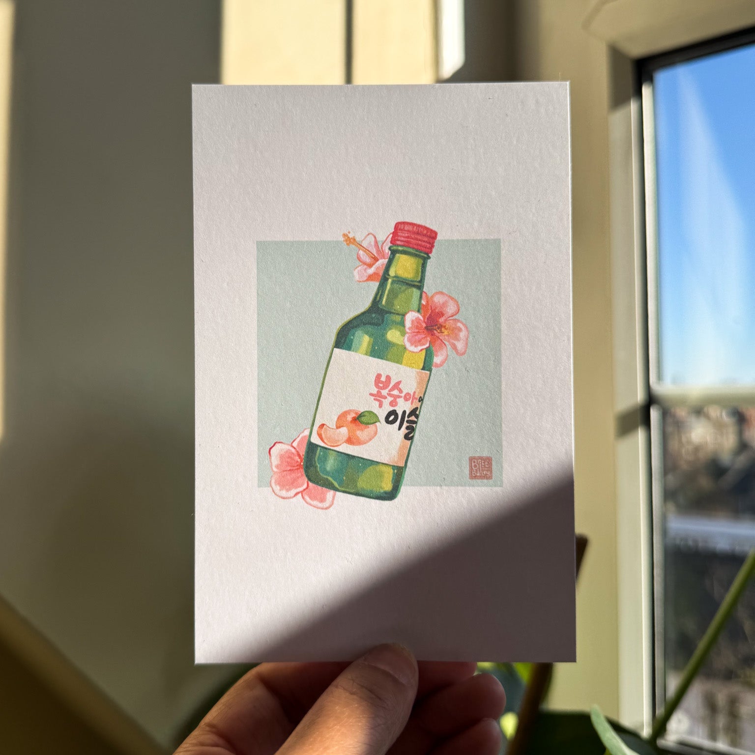 Hand holding a postcard featuring a painterly digital illustration of a bottle of peach flavoured korean soju surrounded by pink hibiscus flowers on a soft teal background. 