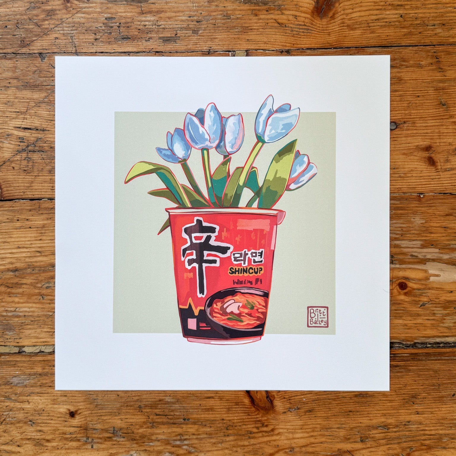 Hand holding an art print featuring a painterly digital illustration of cup of shin ramen noodles with a bouquet of white tulips inside on a soft green background. 