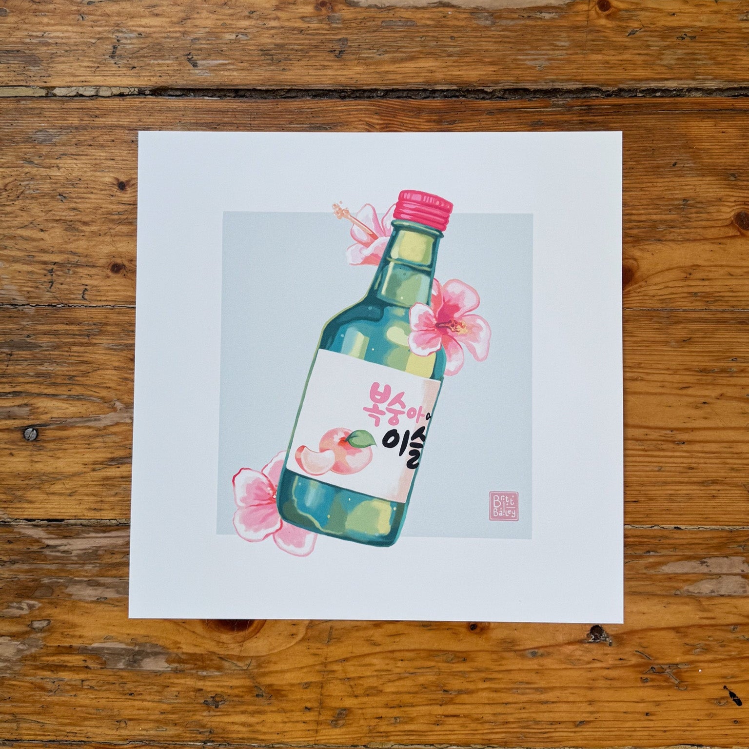 Hand holding an art print featuring a painterly digital illustration of a bottle of peach flavoured korean soju surrounded by pink hibiscus flowers on a soft teal background. 