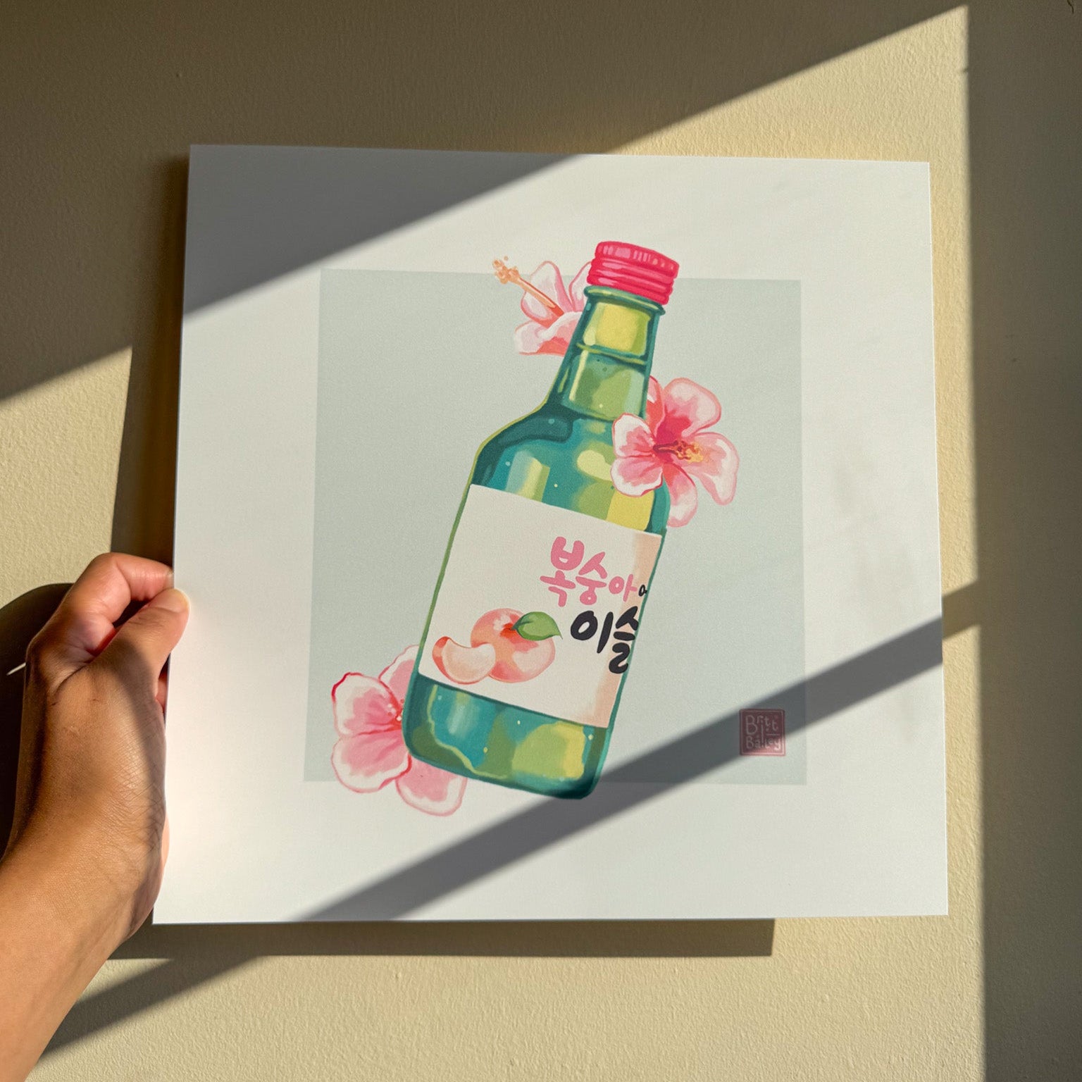 Hand holding an art print featuring a painterly digital illustration of a bottle of peach flavoured korean soju surrounded by pink hibiscus flowers on a soft teal background. 