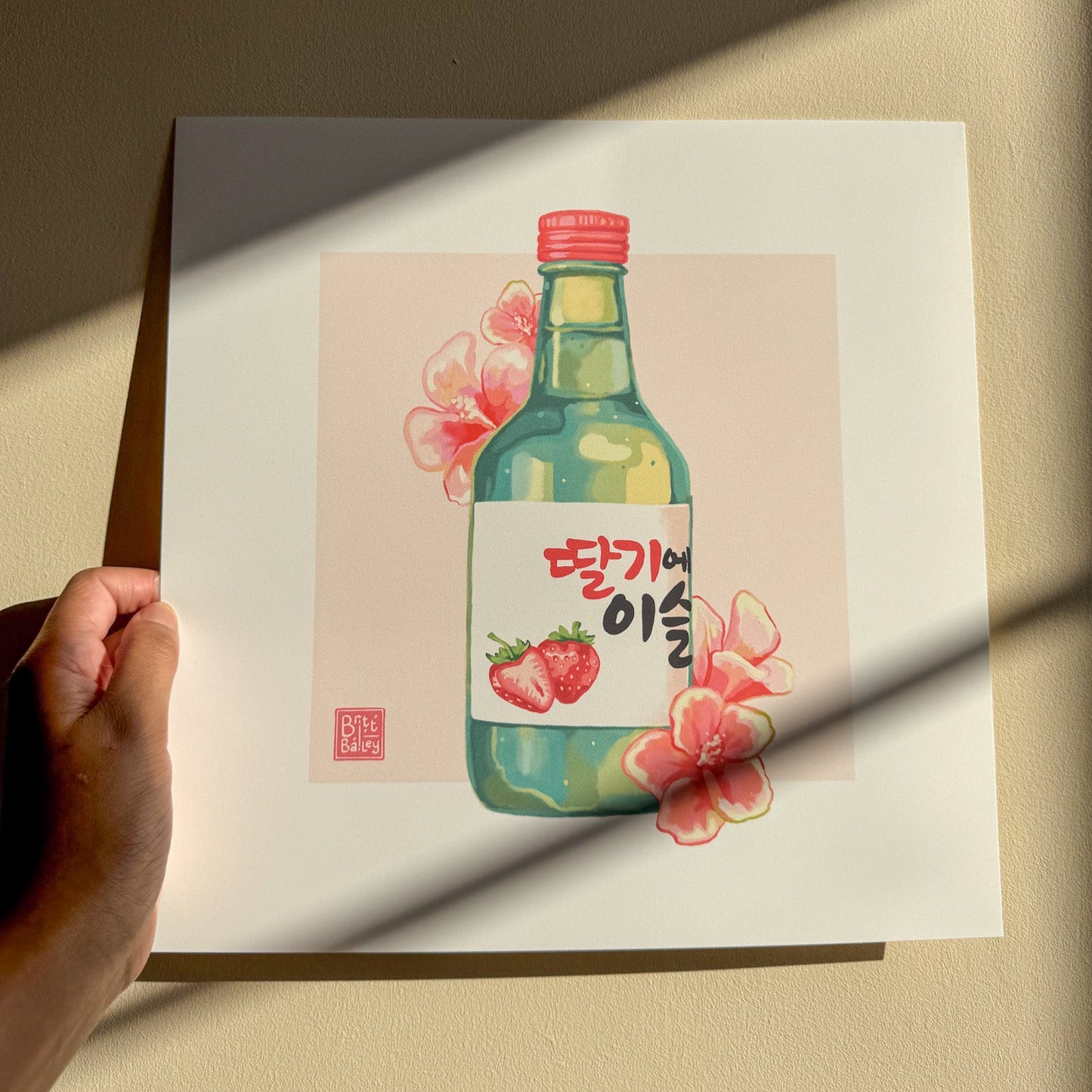 Hand holding an art print featuring a painterly digital illustration of a bottle of strawberry flavoured korean soju surrounded by red hibiscus flowers on a soft pink background. 