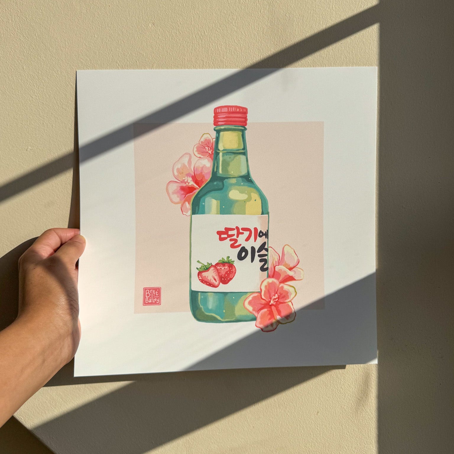 Hand holding an art print featuring a painterly digital illustration of a bottle of strawberry flavoured korean soju surrounded by red hibiscus flowers on a soft pink background. 
