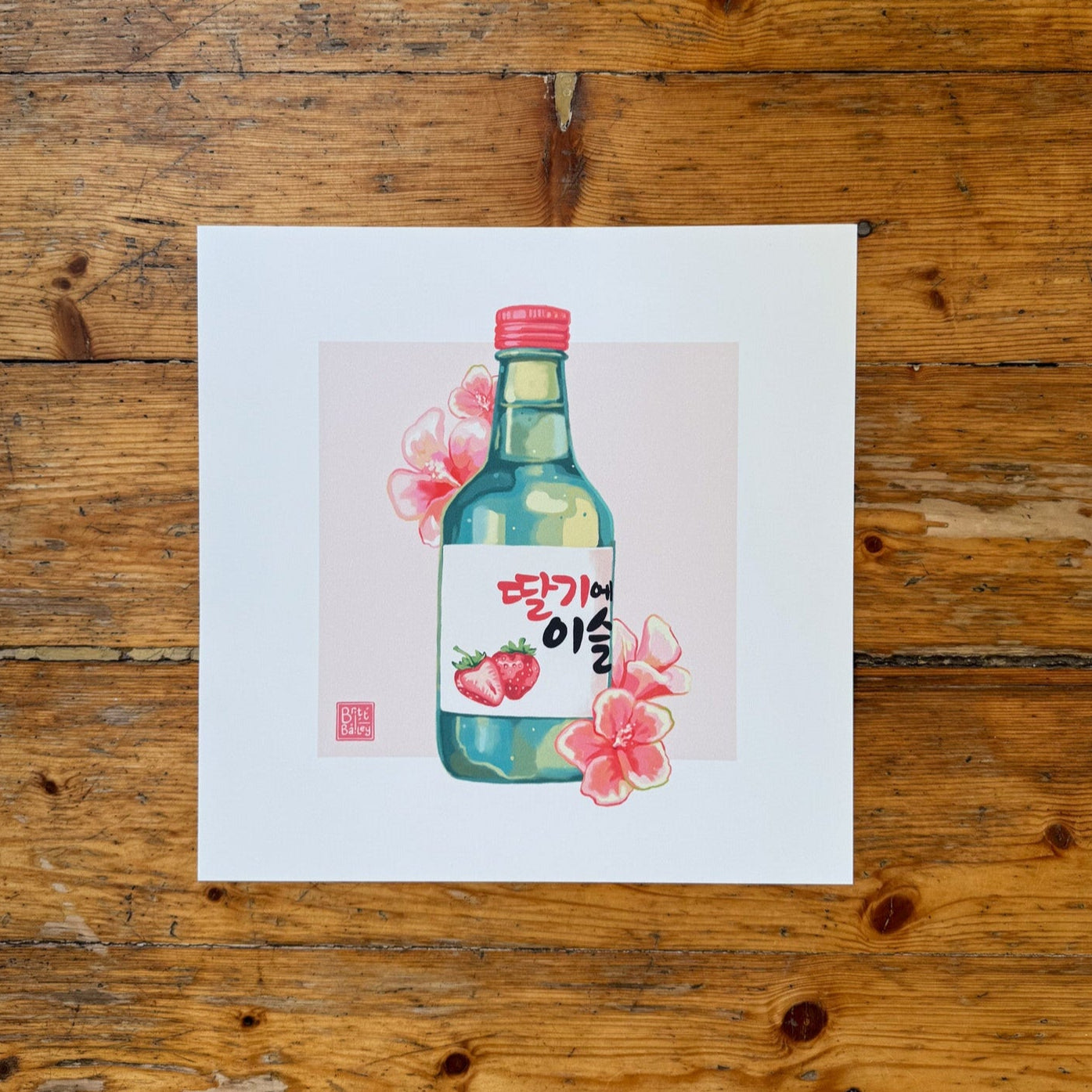 Hand holding an art print featuring a painterly digital illustration of a bottle of strawberry flavoured korean soju surrounded by red hibiscus flowers on a soft pink background. 